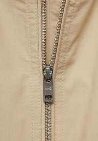 Beige stof med en metallisk lynlås, der har et robust kæde-link design. Lynlåsens træk viser brandets logo "NKO."