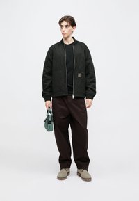 Μαύρο μπουφάν bomber με ραμμένη επένδυση και ριμπ μανσέτες, φορεμένο πάνω από μαύρο πουκάμισο. Φαρδιά καφέ παντελόνια και γκρι μποτάκια. Κρατάει μια μικρή πράσινη τσάντα.