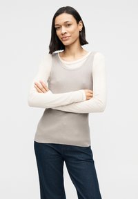 Canotta grigia a coste indossata sopra una maglia crema a maniche lunghe, abbinata a pantaloni blu scuro. Tessuto liscio, scollo a U e design aderente.