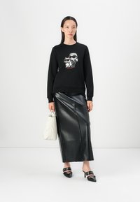 Felpa nera con stampa grafica, abbinata a una maxi gonna in pelle nera lucida. Il modello tiene in mano una borsa a tracolla bianca trapuntata. Sandali neri con tacco alto.