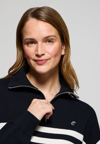 Marinblå zip-tröja med vita ränder, ribbade muddar och en liten logotyp på bröstet. Mjuk textur och något strukturerad design.