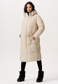 Beige polstret lang jakke med en t�ætsiddende hætte, lynlås lukning og sidelommer. Båret over sorte leggings og pelsforede ankelstøvler.