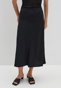 Schwarzer Midi-Rock aus glattem Stoff mit hoher Taille, fließender Silhouette und subtiler Textur. Kombiniert mit klobigen schwarzen Riemchensandalen.