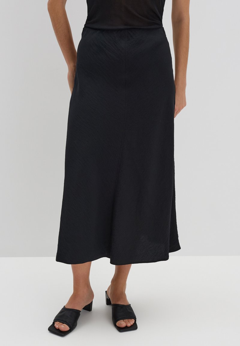 Schwarzer Midi-Rock aus glattem Stoff mit hoher Taille, fließender Silhouette und subtiler Textur. Kombiniert mit klobigen schwarzen Riemchensandalen.