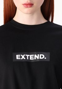 Melnas kokvilnas T-krekls ar taisnstūrveida grafiku, kurā centrā uz krūtīm ir biezajās baltajās burtiem rakstīts "EXTEND."