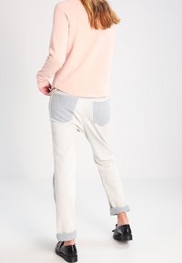 Suéter rosa claro con dobladillo redondeado, combinado con pantalones de mezclilla en un tono gris claro y blanco que cuentan con dos bolsillos de parche grises. Se ven zapatos negros.
