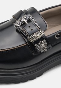 Svart skinnloafers med en dekorativ metallspenne med intrikat detaljer og en kontrasterende teksturert gummisåle.