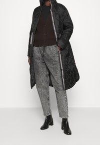 Abrigo largo acolchado negro con forro a rayas en contraste, llevado sobre un cárdigan marrón y pantalones grises texturizados, combinado con botines negros de tacón.