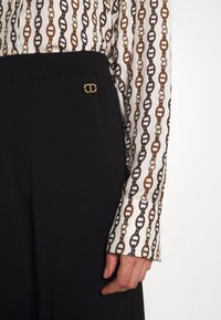 Blouse blanche avec motif de chaîne en marron et or, associée à un pantalon noir avec une taille large et un détail doré.