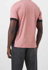 Muž v růžovém polo tričku s černými bočními panely a tmavě šedých džínách, zobrazený zezadu na jednobarevném pozadí.