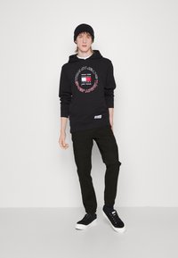 Zwart hoodie met een circulair logo-ontwerp, gecombineerd met donkere cargo-broeken en zwarte sneakers. Model draagt een zwarte muts en staat tegen een witte achtergrond.