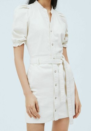 Woman wearing a cream button-front dress with puff sleeves and a fabric belt tied at the waist, standing against a plain background.