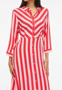 Femme portant une robe chemise à rayures verticales rouges et rose clair, manches 3/4 et col, debout les mains le long du corps.