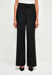 Pantalons larges noirs en tissu texturé, dotés de deux poches avant, d'un devant plat et d'une taille ajustée. Talons noirs portés.