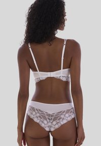 Mujer con cabello rizado que lleva un sujetador de encaje blanco y unas braguitas de encaje a juego, mostrada de espaldas contra un fondo gris liso.