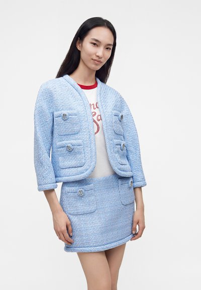 Femme portant un ensemble veste et jupe bleu clair texturé avec poches sur une chemise blanche avec texte rouge, debout devant un fond uni.