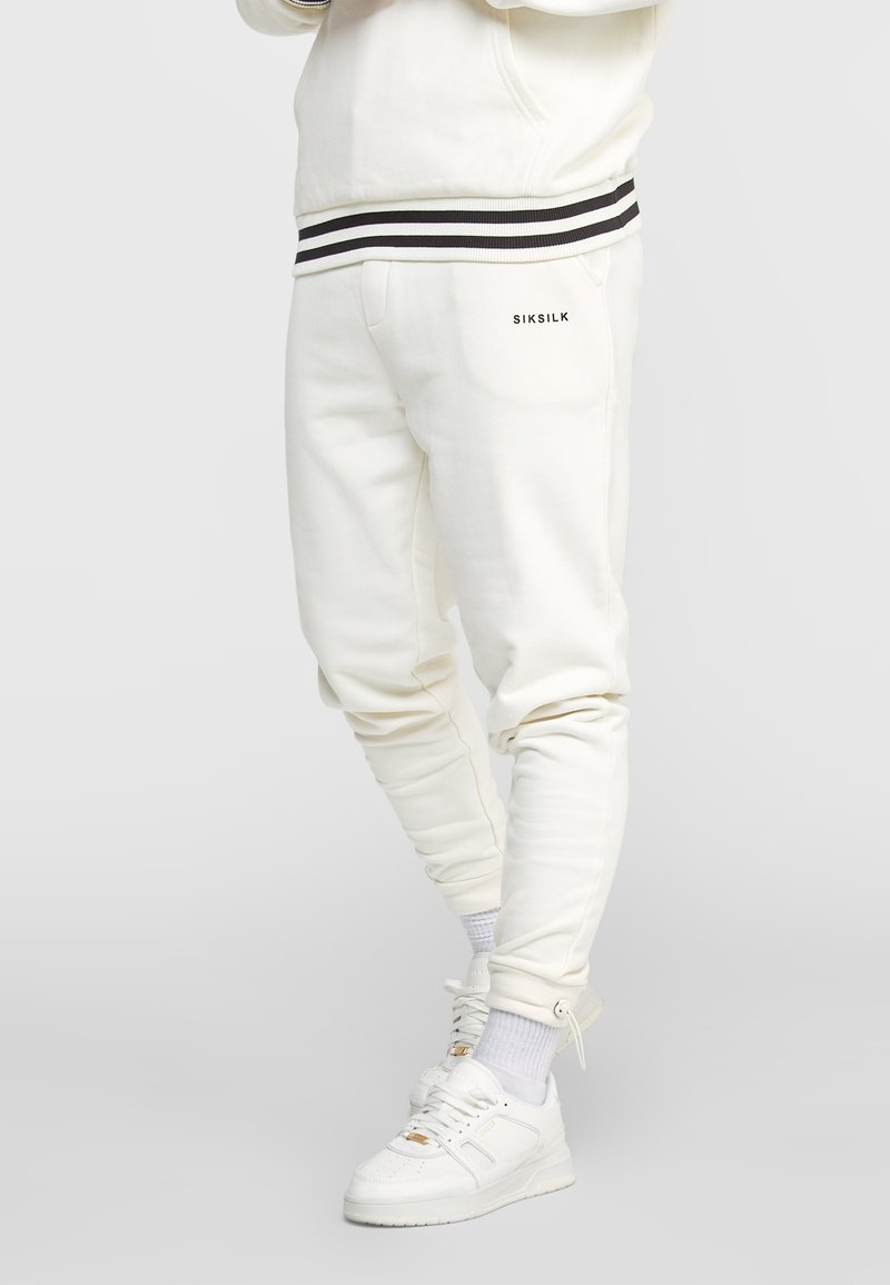 Jogging crème à coupe slim, avec poignets côtelés et taille élastique. Accents à rayures noires et blanches et détail de poche latérale.