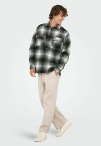 Camisa de franela a cuadros en negro, blanco y gris, con dos bolsillos en el pecho y cierre de botones; modelo lleva pantalones beige claros y zapatillas.