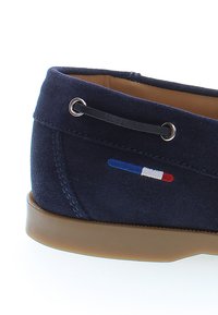 Navyblauer Wildleder-Schlüpfschuh mit brauner Sohle, schwarzen Schnürsenkeln durch Metallösen und einem kleinen horizontalen Trikolore-Detail in Blau, Weiß und Rot.
