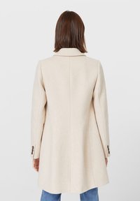 Manteau en laine beige au design structuré, col cranté et manches longues. Comprend des poignets boutonnés et descend sous les hanches.