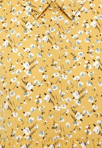 Żółta tkanina w kwiaty o teksturowej powierzchni. Posiada rozproszone białe i niebieskie kwiaty, czarne kontury oraz kołnierzyk z guzikami.