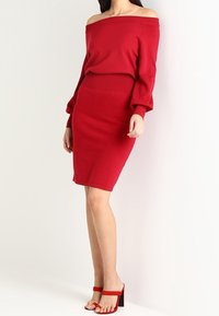 Maglione rosso lavorato a maglia con scollo off-shoulder abbinato a una gonna a tubino aderente, con maniche lunghe e orlo a costine. Eleganti sandali rossi con tacco alto.