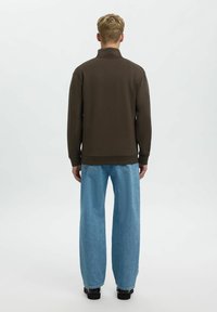 Hnědá mikina s vysokým límcem, kombinovaná s volnými světle modrými džínami. Materiál se jeví jako měkký, se silnými švy a bez viditelných log.