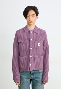 Lila Strickjacke mit Kragen, geripptem Muster, zwei vorderen Taschen und weißen Knöpfen, kombiniert mit hellblauen Jeans.