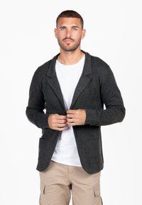 Blazer in lana grigio scuro con revers, tasche frontali e chiusura a bottone, indossato sopra una maglietta bianca e pantaloni beige chiaro.