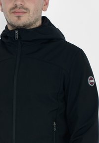 Giacca softshell nera con zip frontale, cappuccio regolabile e tasche laterali. Presenta un patch con logo sulla spalla sinistra.