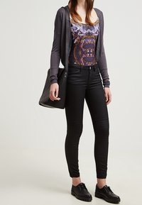 Svarta skinny jeans med texturerat ribbat detaljer, matchat med en mönstrad tank top och en grå cardigan. Svarta skor och en svart axelväska finns närvarande.