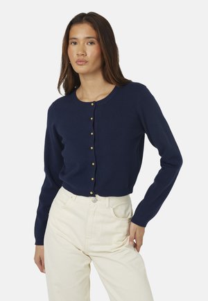 Vrouw draagt een marineblauwe, met goudkleurige knopen gebuttonede cardigan en crèmekleurige broek met hoge taille, staand met één hand op de heup.