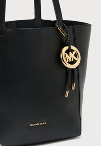 Sac fourre-tout en cuir noir avec deux poignées, orné d'un médaillon rond à logo doré et d'un accent métallique. Nom de la marque en relief doré sur le devant.