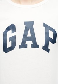 Zone de la poitrine d'une chemise blanche avec de grandes lettres bleues marines épaisses formant le mot "GAP" à l'avant.