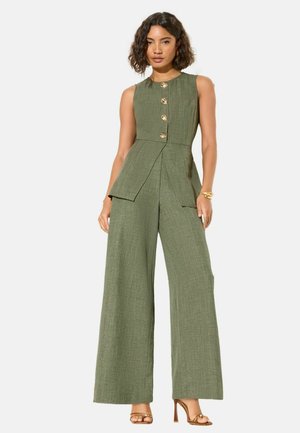 Nő ujjatlan olívazöld overált visel, amely bő szárú és arany gombokkal díszített, arany magassarkú szandállal és karkötővel párosítva.