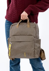 Ruskea teksturoidun pinnan reppu, jossa on kaksi yläkahvaa, etutasku, keltainen vetoketjun vedin ja säädettävä olkahihna. Siinä on minimalistinen muotoilu.