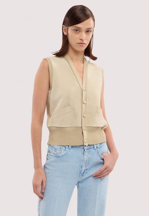 Donna che indossa un gilet beige senza maniche con bottoni e jeans azzurri, in piedi contro uno sfondo chiaro uniforme con una mano in tasca.