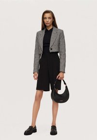 Czarno-biała krótka marynarka w wzór pepitki, czarny top, dopasowane czarne szorty, czarne mokasyny, trzyma gładką czarną torebkę.