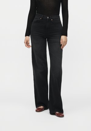 Person iført sorte højtaljede, brede jeans, en sort langærmet top og brune sko, stående mod en ensfarvet grå baggrund.