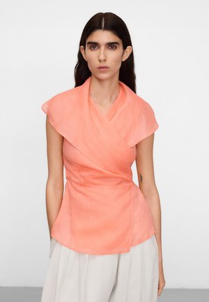 Femme aux longs cheveux foncés portant un haut sans manches corail de style cache-cœur et un pantalon beige clair à larges jambes, debout devant un fond uni.