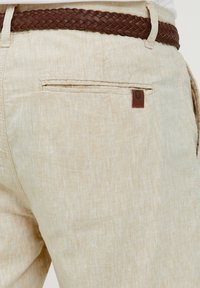 Lysbeige linnedragt med en tekstureret overflade, der har et brunt flettet bælte og et lille brunt logo-tag over baglommen.