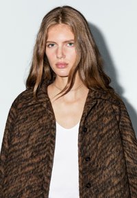 Manteau en laine texturée marron avec un col cranté et des boutons noirs, superposé sur un haut blanc. Motif audacieux et finition douce.