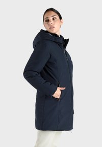 Lange donkerblauwe pufferjas met capuchon, rits aan de voorkant en zijzakken. Glad textuur, lichtgewicht materiaal en moderne snit.