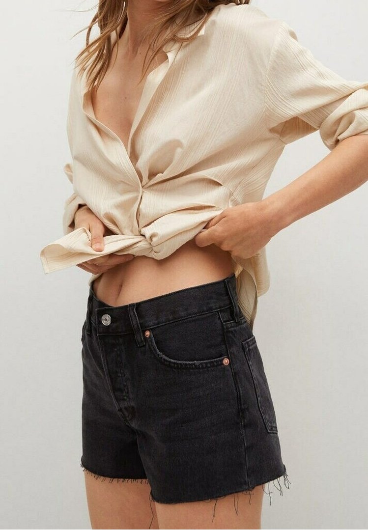 Camisa a rayas beige, anudada en la cintura, combinada con pantalones cortos de mezclilla negros de cintura alta con dobladillos deshilachados y detalles en color cobre.