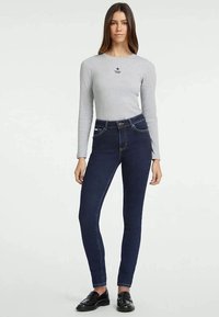 Camisa cinza de mangas longas com um pequeno logótipo de estrela preta; jeans skinny de cintura alta de cor azul-escuro com costuras visíveis e bolsos frontais.