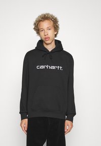 Svart hettegenser i bomull med frontlomme og hvit logo på brystet. Modellen har på seg svarte cordbukser.