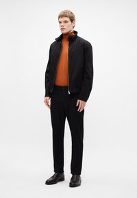 Čierna bomber bunda s postaveným golierom, spárovaná s oranžovým rolákom a čiernymi nohavicami. Hladké textúry, minimalistický dizajn. Čierne topánky.