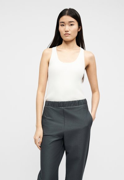 Γυναίκα με μακριά μαύρα μαλλιά, φορώντας λευκό μπλουζάκι χωρίς μανίκια και σκούρο γκρι παντελόνι με ελαστική μέση, στέκεται μπροστά σε απλό φωτεινό φόντο.