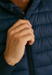 Námořnická modrá prošívaná péřová bunda se zipem vpředu. Zblízka pohled na ruku držící zip. Měkká struktura a lehký design.