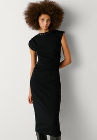 Abito midi aderente nero con un tessuto texturizzato, maniche a cappuccio e dettagli metallici decorativi sulla spalla. Silhouette liscia con drappeggio sottile.